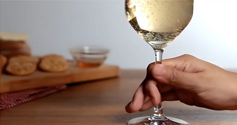 Một bàn tay dùng ngón cái và ngón trỏ kẹp vào phần thân ly, ngay điểm giao với đế ly rượu vang trắng với nền phía sau là bánh quy và mật ong.